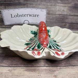 May include: A cream-colored ceramic serving dish shaped like a leaf with a three-dimensional red lobster figure in the center. The lobster is detailed with black spots and green accents. A white sign behind the dish displays the text Lobsterware.