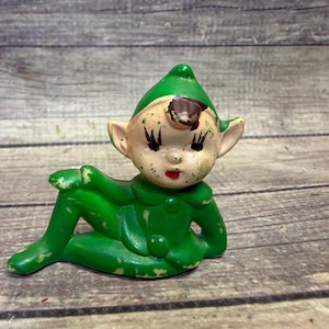 May include: A vintage ceramic elf figurine in a reclining pose. The elf is primarily green with a pointed hat, rosy cheeks, and a red mouth. The figurine has visible wear and tear, adding to its antique charm.