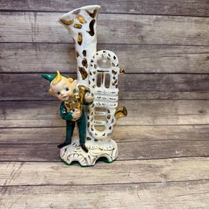 May include: A vintage ceramic vase featuring a whimsical elf figure. The vase is white with gold accents and a trumpet-like shape. The elf is dressed in green and holds a golden instrument, adding a touch of fantasy to the decorative piece.