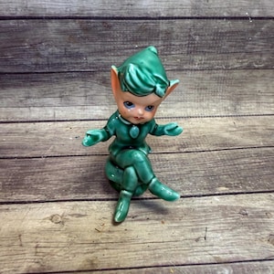 May include: A ceramic figurine of a seated elf with green clothing and a matching pointed hat. The elf has large, pointed ears, blue eyes, and is posed with arms outstretched. The figurine is set against a weathered wooden background.