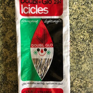 May include: A package of Double Glo Icicles, a vintage Christmas decoration. The package is white with red and green accents, and features the text "Double Glo Icicles" and "150 strands, 18 inches long". The design includes a stylized face.