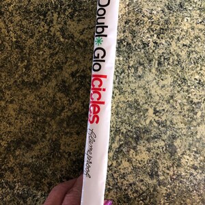 May include: A white package with the words "Double Glo Icicles" printed in black and red. A green asterisk is between "Double" and "Glo". The text "Flameproof" is written in cursive. The package is held by a person.