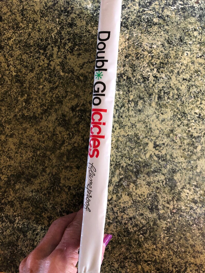May include: A white package with the words "Double Glo Icicles" printed in black and red. A green asterisk is between "Double" and "Glo". The text "Flameproof" is written in cursive. The package is held by a person.