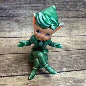 May include: A vintage ceramic elf figurine in a seated pose. The elf is predominantly green with a pointed hat, rosy cheeks, and blue eyes. The figurine has outstretched arms and crossed legs, with a glossy finish.