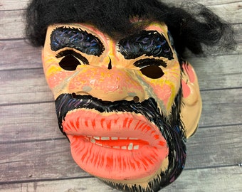 May include: A vintage plastic Halloween mask with a black wig, painted eyebrows, a mustache, and a large, open mouth with pink lips and white teeth.