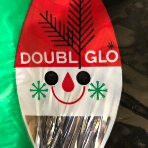 May include: A vintage Christmas ornament package with a red and white design. The package features the text "DOUBLE GLO", a black tree graphic, and a smiling face with green star accents. Silver tinsel hangs from the bottom.