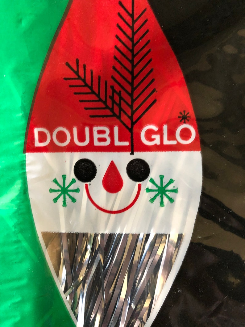 May include: A vintage Christmas ornament package with a red and white design. The package features the text "DOUBLE GLO", a black tree graphic, and a smiling face with green star accents. Silver tinsel hangs from the bottom.