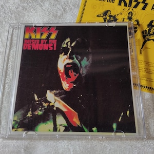 May include: A CD case featuring the album cover for "Raised by the Demons!" by the band KISS. The cover art depicts a close-up of a band member with open mouth. The KISS logo is at the top. A yellow promotional insert is visible.