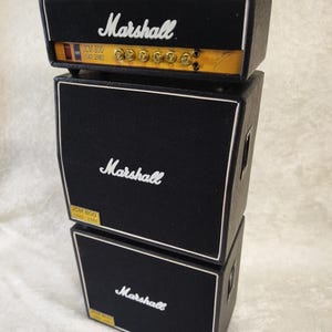 Könnte beinhalten: Ein Stapel von drei schwarzen Marshall-Verstärkern. Der obere Verstärker trägt den Schriftzug "Marshall" in weißer Schrift und einen Griff. Die mittleren und unteren Verstärker tragen ebenfalls den Schriftzug "Marshall". Der obere Verstärker trägt den Schriftzug "JCM 800 LEAD SERIES".