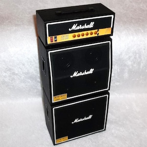 Könnte beinhalten: Ein schwarzer Marshall-Verstärkerstapel mit weißer Umrandung und dem Marshall-Logo. Das obere Gerät trägt den Text "JCM 800 LEAD SERIES". Der Stapel umfasst einen Verstärkerkopf und zwei Lautsprecherboxen, ein klassisches Design für Gitarrenverstärkung.