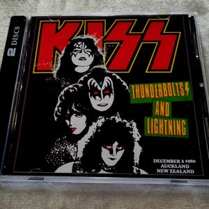 May include: A CD case featuring the band KISS. The cover art displays the band members' faces in makeup, with the band's name in red and the album title "Thunderbolts and Lightning" in green. The text "2 DISCS" is on the side.