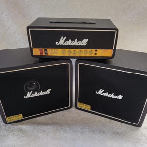 May include: Three black Marshall amplifiers. The top amplifier has a handle and a yellow stripe with the text "JCM 800 LEAD SERIES". The other two amplifiers have the Marshall logo in white.
