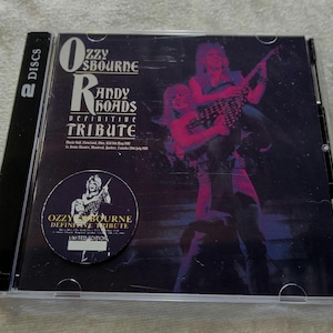 May include: A CD case for the Ozzy Osbourne Randy Rhoads Definitive Tribute album. The cover features a purple and pink image of musicians playing guitars. A circular sticker on the front reads "Ozzy Osbourne Definitive Tribute Limited Edition."
