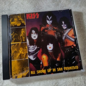 Puede incluir: Una caja de CD con la banda KISS. La portada del álbum tiene un fondo oscuro con los miembros de la banda con maquillaje y disfraces completos. El texto "KISS" es rojo, y el título del álbum "ALL SHOOK UP IN SAN FRANCISCO" es visible.