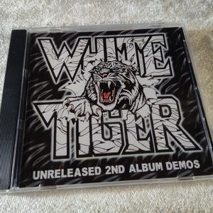 May include: A CD case featuring the album "WHITE TIGER" with a black and white tiger graphic. The text "UNRELEASED 2ND ALBUM DEMOS" is below the title. The case is clear with a black spine.