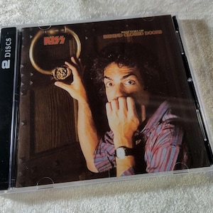 May include: A CD case featuring the album cover for "What Lives on Behind Closed Doors" by KISS. The cover art shows a man with a surprised expression, reaching for a door knocker. The case is labeled "2 DISCS".