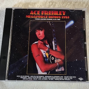 Peut inclure: Boîtier de CD avec l'album "Megaforce Demos 1985" d'Ace Frehley. La pochette présente un portrait d'Ace Frehley tenant une guitare sur fond étoilé. La liste des titres est en bas. Le texte "ACE FREHLEY" est en haut.