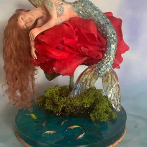 May include: A handcrafted mermaid sculpture with long red hair, resting on a large red rose. The mermaid has a sparkling blue tail and wings. The base features a pond with small fish and green moss.