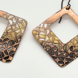 May include: A pair of copper-colored earrings with a geometric design. The earrings are diamond-shaped with a cutout center and feature a pattern of small, semi-circular shapes in shades of white, yellow, and black. The ear wires are dark metal.