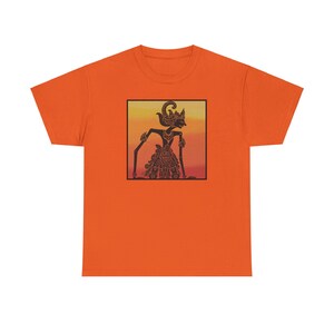 Puede incluir: Camiseta naranja con una ilustración en negro y dorado de una marioneta Wayang Kulit. La marioneta está situada sobre un fondo cuadrado con un degradado de amarillo, naranja y rojo. La camiseta es de color naranja liso.