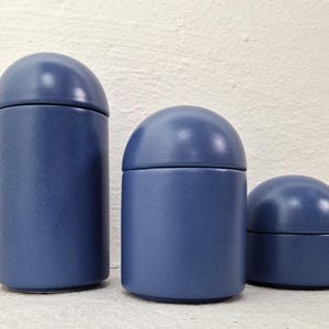 Pode incluir: Três potes de cerâmica azuis com tampas arredondadas. Os potes são cilíndricos e variam em tamanho, com o pote maior à esquerda e o menor à direita. Os potes são de cor azul fosco e estão sobre uma superfície branca.