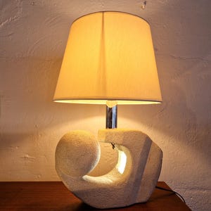 May include: A white ceramic table lamp with an abstract, sculpted base and a white fabric lampshade. The lampshade is lit and casts a warm glow.