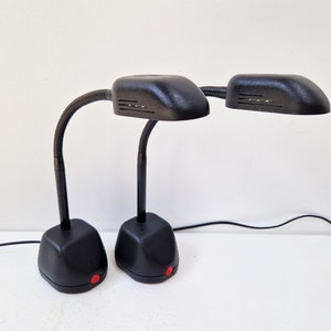 Può includere: Due lampade da scrivania nere con bracci flessibili a collo di cigno e paralumi rettangolari. Ogni lampada ha una base quadrata con un pulsante di accensione rosso. Le lampade sono collegate a una presa di corrente con un cavo nero.