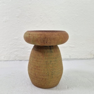 Può includere: Un oggetto decorativo fatto a mano a forma di fungo. Il pezzo presenta una parte superiore arrotondata e una base cilindrica strutturata. Il colore è una miscela di toni terrosi, con una parte superiore marrone chiaro e una base marrone verdastra. L'oggetto è realizzato con un materiale naturale.