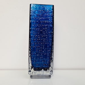 May include: A tall, rectangular blue glass vase with a textured surface. The vase has a clear glass base.