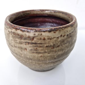 Könnte beinhalten: Eine kleine, runde Keramikschale mit hellbrauner Glasur und einem dunkelbraunen Rand. Die Schale hat eine strukturierte Oberfläche und scheint handgefertigt zu sein.
