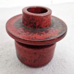 Może przedstawiać: Czerwony ceramiczny świecznik z okrągłą podstawą i cylindrycznym wierzchem. Świecznik ma teksturowaną powierzchnię z ciemnobrązowymi plamkami. Design jest prosty i nowoczesny, odpowiedni do różnych stylów wystroju wnętrz.