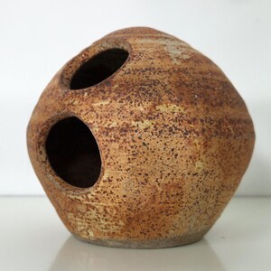 Op de afbeelding: Een bolvormig keramieken vat met een gespikkeld beige en bruin glazuur. Het object heeft twee ronde openingen aan de bovenzijde en toont een getextureerde, aardse afwerking met donkerbruine vlekken over het ronde, met de hand gevormde kleilichaam.