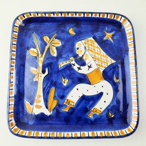 Peut inclure: Une assiette carrée en céramique bleue avec un motif peint à la main représentant une personne jouant de la flûte devant un arbre. Le motif est dans des tons de jaune, blanc et orange sur un fond bleu.