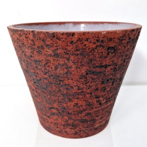 Könnte beinhalten: Ein terracottafarbener Keramiktopf mit einer rauen, strukturierten Oberfläche. Der Topf hat eine breite, abgerundete Oberseite und eine leicht konische Basis.