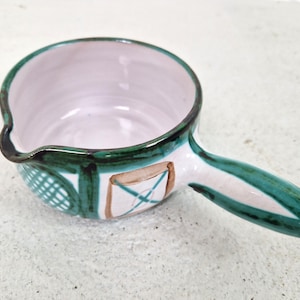 Op de afbeelding: Handbeschilderde keramische sauskom met een handvat. De kom heeft een wit interieur en een groen en wit exterieur ontwerp. De handgreep is groen en wit. De kom heeft een tuit om gemakkelijk te schenken. De kom heeft een decoratief ontwerp aan de zijkant.