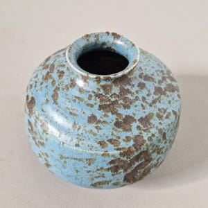 Puede incluir: Un pequeño jarrón de cerámica redondo con un cuerpo ancho y achaparrado y una abertura circular estrecha en la parte superior. La superficie presenta un esmalte base azul claro con un patrón moteado de tonos marrón oscuro y tierra.