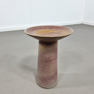 May include: A pedestal bird bath with a wide, shallow bowl and a cylindrical base. The bird bath is a speckled, muted reddish-purple color with a yellow-gold interior. The design is simple and modern, suitable for a garden or patio.