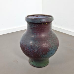 Può includere: Un vaso in ceramica con corpo bulboso e bordo largo e svasato. La superficie presenta una smaltatura maculata con toni bordeaux profondo, blu scuro e verde bosco, creando un aspetto strutturato e punteggiato su tutto l'esterno del recipiente.