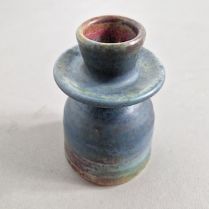 Pode incluir: Um vaso de cerâmica feito à mão com um design único. O vaso apresenta um esmalte azul com toques de verde e castanho. A parte superior tem uma pequena abertura com um interior castanho avermelhado e uma placa circular. Uma peça decorativa.