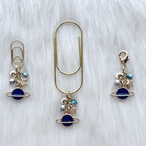 May include: Three gold-colored paper clips and charms on a white fluffy surface. Each charm features a blue planet design with a gold ring, surrounded by small silver and gold stars and beads. The paper clips vary in size.
