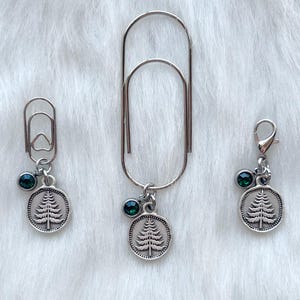 May include: Three silver-toned paper clips with decorative charms. Each charm features a tree design within a circular frame and a small, dark green gem. The paper clips are arranged on a white, fluffy surface.