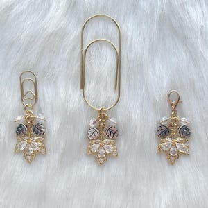 May include: Three gold-colored paper clips with leaf-shaped charms. The charms feature clear crystals, small silver and gray leaves, and small clear beads. The paper clips are arranged on a white, fluffy surface.