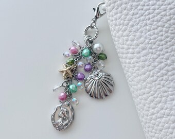 May include: A mermaid-themed charm with a lobster clasp. The charm features a silver seashell, starfish, and mermaid pendant, along with colorful beads in shades of pink, green, purple, and clear. The charm is attached to a white leather item.