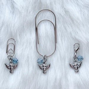 May include: Three silver-toned paper clips with decorative charms. Each charm features a small owl pendant with outstretched wings and a cluster of light blue beads. The paper clips are arranged on a white, fluffy surface.
