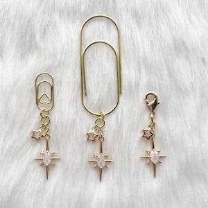May include: Gold-coloured paper clips with star and crystal charms. The charms feature a small star and a larger starburst design with a central crystal. The paper clips are in different sizes, one with a clasp.