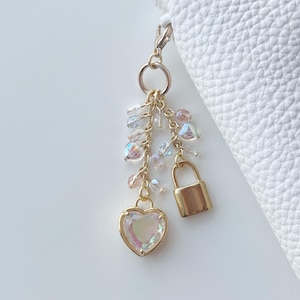May include: A gold-toned keychain charm featuring a heart-shaped iridescent crystal pendant, a small gold padlock, and dangling clear and pink beads. The charm is attached to a gold clasp, with a white leather item in the background.