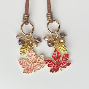 May include: Two decorative charms with maple leaf pendants. Each charm features a gold ring, brown suede cord, and a cluster of beads and a gold pinecone. One leaf is pink, the other is red.