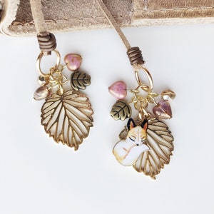 May include: Two decorative bookmark charms with tan suede cords. Each charm features a gold-colored leaf, pink heart-shaped beads, and a sleeping fox charm. The charms are connected with gold-colored chains and rings.