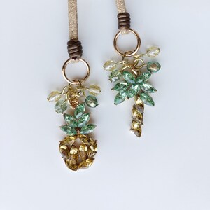 May include: Two gold-toned bag charms with decorative crystal pineapple and floral designs. The charms feature green, yellow, and brown crystals, attached to gold rings and tan cords. The charms are hanging from a light gray bag.