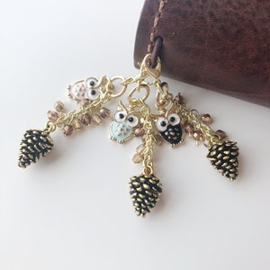 May include: A gold-toned charm with three owl pendants in pink, light blue, and black, accented with small brown beads and gold pinecone charms. The charm is attached to a brown leather item.